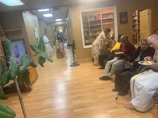 Interior hallway of a temple or ashram with light wood-look flooring and a tall green plant with broad leaves in the left foreground. On the right, several seated people sit along a wall; one older man in a dark jacket and glasses holds a white plate, another in a light-colored top is seated beside him, and a bald man in a light shirt sits at the far right with a white plate in his lap. A robed figure in a light garment stands in the background near shelves of books. A portrait-style picture hangs on the left wall beside a small table. The space appears to be a communal dining or resting area.