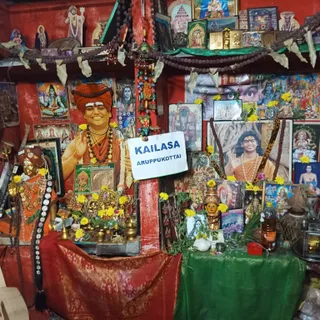 A small shrine room with red painted walls, photographed indoors. A large printed portrait on the left shows a smiling figure in an orange turban, orange robe, rudraksha mala and gold necklace, with one hand raised in blessing; this is a depiction, and the page-based appearance cues are several (turban, orange clothing, rudraksha, ornate necklace), so it is labelled Swamiji (probable). A white paper sign reads KAILASA ARUPPUKOTTAI in blue letters and hangs at the centre. Below and beside it is a green-cloth altar with brass lamps, a kalasha-like vessel, yellow flower garlands and marigolds, a small golden deity murti, and a red-draped table. Framed prints of deities, a Shiva image and other devotional pictures cover the walls, along with rudraksha malas and strings of marigolds. Another framed portrait on the right shows a young figure in saffron robes with a raised hand and pearl mala. No living people are visible.