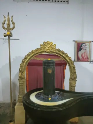 A photograph of a dark cylindrical Shiva linga standing on a black yoni pedestal with a white border, decorated with yellow marigold flowers and a red tilak mark on its side. Behind it is an ornate gold arched frame or torana with a red and orange cloth canopy. To the left stands a tall gold trident (trishul) with a red mark. On the right wall hangs a framed printed portrait labelled with no readable caption; the man pictured has long dark hair, a rudraksha mala and a red-orange robe, smiling, which is a probable Swamiji depiction. No physical people are present in the scene.