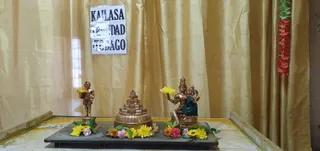 A small altar display on a dark wooden tray set on a tiled floor in front of gold curtains. A paper sign reading KAILASA TRINIDAD & TOBAGO, partly torn and taped, hangs at the top centre. At the centre stands a golden tiered pyramid-shaped sri chakra-style meru with a red dot on its summit. To the left stands a small golden statue of a standing male devotee holding a yellow flower. To the right sits a golden deity murti with a crown, seated with a green cloth draped over its lap and holding a yellow flower, with a small figure beside it. Yellow chrysanthemums, orange and pink flowers and green leaves are arranged around the base. A red and green flower garland hangs at the right edge. No living people are visible.