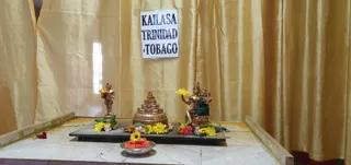 A shrine arrangement in front of gold curtains with a paper sign reading KAILASA TRINIDAD & TOBAGO taped at the centre top. On a dark slate platform sit a small bronze standing figure holding a yellow flower on the left, a tiered bronze pyramid-shaped sculpture in the centre, and a bronze seated deity murti with multiple arms on the right, draped in green cloth and holding a yellow flower. Yellow, orange, pink and white flowers are scattered across the platform, and a steel bowl of cut fruit sits on the floor in front. No people are visible.