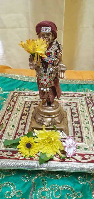 A small bronze statue of a standing figure in a maroon turban with a jewelled forehead ornament, wearing a maroon robe and layered jewelled necklaces with red, green and white stones and pearls. The statue holds a yellow flower in one raised hand and stands on a lotus-shaped base placed on a gold-bordered red and green cloth. Yellow chrysanthemum-style flowers and a pink carnation lie at its feet, with a yellow curtain behind. The scene is a decorated puja display of the statue, with no physical people present.