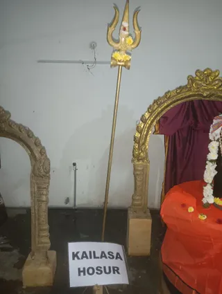 A photograph inside a temple hall showing a tall golden trident (trishul) standing upright on a stand, its prongs decorated with yellow flowers and a red-and-white tilak mark. Beside it are two ornate gold-toned arched frames carved with floral and scrollwork motifs on stone bases. On the right, a deity murti sits draped in a bright orange cloth with a purple curtain behind it, adorned with a white flower garland and yellow marigold petals scattered on the cloth. A white paper sign on the floor reads KAILASA HOSUR. No people are visible.