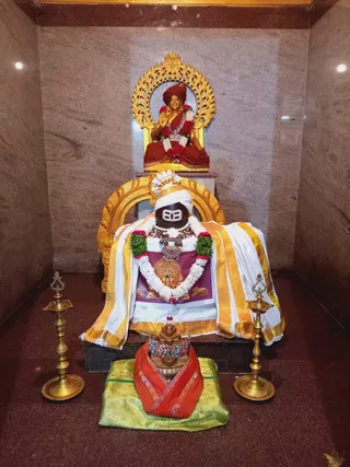 Shrine niche in a polished granite-walled room showing a black stone Shiva lingam draped with white and gold-bordered cloth and garlanded with white and green flower garlands, with a rudraksha-style necklace and a gold-framed portrait-like panel at its chest. Behind it, a golden arched frame holds a gilded statue of a seated saint in red robes and red turban, hands raised in blessing, with a red and white garland. A pair of brass oil lamps stands on either side, and in front a small ornate red-and-gold draped object sits on a green brocade cloth. No physical people are visible; the statues and deity murti are depictions.