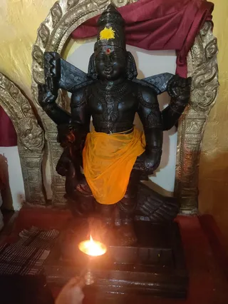 A photograph of a dark stone deity murti standing in a niche framed by ornate golden arch surrounds, with a maroon cloth draped behind. The murti wears a tall crown with a yellow flower, a red tilak on the forehead, and an orange-yellow dhoti, and holds attributes in its raised hands. In the foreground, a small oil lamp burns on a dark stone pedestal, and a hand holding a lit match or stick is partly visible at the lower edge. A folded striped cloth lies to the left. No physical people are fully visible.