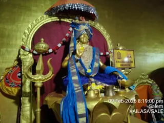 A golden statue of a seated figure in a gilded throne-like alcove with a red canopy above. The figure wears a blue and gold-bordered shawl, blue garment, gold necklaces and a blue headdress, with hands joined in a namaskara gesture. A golden trident stands to the left, garlanded with red and white flowers, and brass vessels, a bell and a kalasha sit before the figure with yellow flowers. A small framed sign at right reads KAILASA SALEM BHARATHI. The timestamp reads 09-Feb-2026 8:19:00 pm.