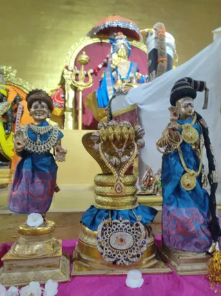 Photo of a temple altar with three decorated figures on gold pedestals against a gold-painted wall. Two standing figures on the left and right wear blue silk sarees with pink and red borders, heavy jewellery, and garlands. The central figure is a cobra-hooded golden deity form with a jewelled necklace and a large jewelled pendant at its base. White flowers lie on a magenta cloth. Behind them stands a gilded trident, a red-and-gold umbrella with sequins, and a deity image draped in blue cloth. No physical people are visible.
