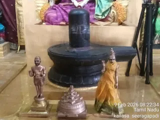 A photograph of a black stone Shivalinga on a black yoni-shaped peetha inside a shrine, with a purple and gold draped figure and garland visible behind it. In front stand two small bronze figures: a standing male figure with a raised hand on the left and a female figure in a yellow sari with a crown on the right, with a bronze Sri Yantra-style meru base between them. A tripod stand and a green cloth appear at the right edge. The camera timestamp reads 9 Feb 2026 08:22:34, Tamil Nadu, kailasa seeragapadi.