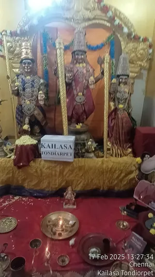Indoor shrine photo with a gold-framed arch backdrop decorated with flower garlands. Three deity murti stand on a raised golden cloth platform: the figure on the left wears a blue dress with a crown and silver-and-gold garlands, the central figure wears a red-pink sari with a tall silver crown and a garland, and the figure on the right wears a red sari with a tall silver crown and a garland. A white rectangular sign reads KAILASA TONDIARPET. In front, a red cloth table holds brass plates, a brass lamp with a lit oil flame, small brass bowls and a conch-shaped offering, with a small figure and yellow flowers nearby. No physical people are visible.