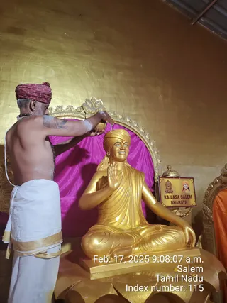 A photograph of a priest performing abhishekam on a gold-painted statue seated in lotus pose with the right hand raised in blessing and a golden turban on its head. The priest is bare-chested with a white dhoti bordered in gold, wears a red patterned turban and a rudraksha mala, and pours liquid from a small vessel onto the statue's head. The statue sits before a magenta drape within a gold ornate arch, beside a small sign reading KAILASA SALEM BHARATH with a portrait image. A timestamp reads Feb. 17, 2025, Salem, Tamil Nadu. The statue is a depiction, not a physical person.