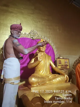 A shirtless priest in a red and gold turban, white dhoti with gold border and a rudraksha mala, performs a ritual by raising a lit oil lamp above the head of a golden seated statue. The statue sits in a meditative posture with one hand raised in blessing, on a lotus base before a magenta drape and ornate golden arch. A placard reading KAILASA SALEM BHARATH with a small portrait sits beside it. A timestamp reads Feb. 17, 2025 9:07:21 a.m. Salem, Tamil Nadu. Only the priest is a physical person; the golden figure is a statue.