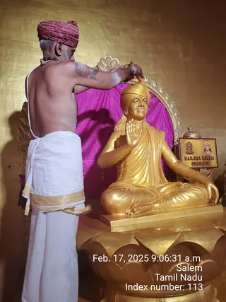A photo of a golden statue of a seated, smiling figure with a golden turban and long hair, in a raised blessing mudra with one hand, set on a golden lotus base against a magenta drape and gilded wall. A bare-chested priest with grey hair, red turban, rudraksha mala and white dhoti with gold border stands behind it, pouring or applying a substance over the statue's head. A small framed sign beside the statue reads KAILASA SALEM BHARATH with small portraits. The timestamp reads Feb. 17, 2025 9:06:31 a.m., Salem, Tamil Nadu.