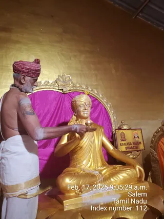A photograph of a priest performing a ritual before a gold-painted seated statue of a saint in meditation pose, wearing a turban with a red bindi and a sash. The priest, bare-chested with grey hair and beard, wears a red-and-gold turban, a rudraksha mala, white dhoti with a gold border, and holds a small oil lamp toward the statue while white paste marks his arm. Behind the statue is a magenta cloth and an ornate gold frame. A small wooden sign on the right reads KAILASA SALEM BHARATH with two small portrait images. A timestamp in the lower corner reads Feb. 17, 2025 9:05:29 a.m. Salem, Tamil Nadu, Index number: 112.