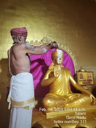 A bare-chested man in a white dhoti with a gold-bordered cloth wrapped around his waist and a red-and-maroon turban stands behind a seated golden statue, applying white paste to its forehead and face with his hands. He wears a rudraksha mala around his neck. The statue sits in lotus pose, one hand raised in blessing, against a gold-leafed wall with a magenta cloth backdrop and an ornate gold arch. A small yellow sign on the right reads KAILASA SALEM BHARATH beside a small portrait of a smiling man in orange, and a brass lamp sits nearby. This is a photo of an abhishekam-style anointing of a statue, and the statue is not a live person.