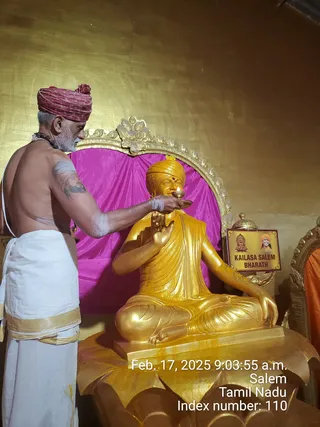 A photograph of a shirtless priest in a red turban and white dhoti with a gold border, holding a small oil lamp with a flame to the face of a seated golden statue. The statue shows a figure in a golden turban with a red tilak, hand raised in blessing and the other hand resting on the lap, seated on a golden lotus base before a magenta drape and an ornate gold arch. A printed sign on the right reads KAILASA SALEM BHARATH beside small pictures of a temple and a saffron-turbaned figure. The priest wears a rudraksha mala and has sacred ash marks on his arm. Timestamp overlay reads Feb. 17, 2025 9:03:55 a.m. Salem, Tamil Nadu. Performing a lamp offering (aarti) to a golden murti.