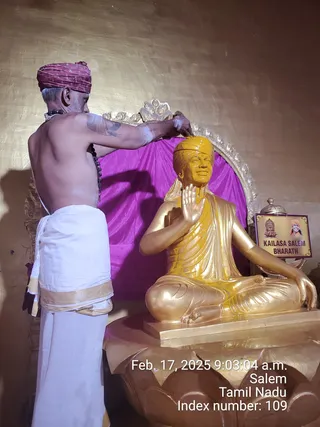 A shirtless priest in a maroon turban, white dhoti with gold border and rudraksha mala stands at left, pouring yellow liquid over the head of a seated golden statue in a mudra pose with raised right hand. The golden statue wears a yellow turban and draped robe and sits on a lotus pedestal before a magenta cloth and an ornate silver-framed backdrop. A small placard reads KAILASA SALEM BHARATH beside it, showing a temple emblem and a portrait. Timestamp overlay reads Feb. 17, 2025 9:03:04 a.m., Salem, Tamil Nadu. Abhishekam ritual on a statue.