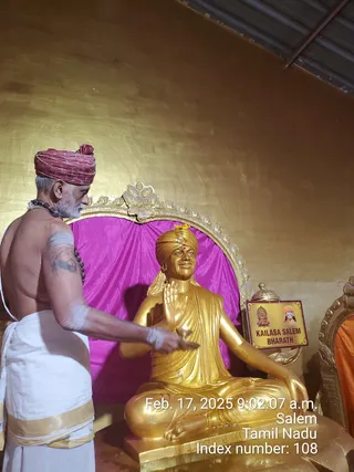 A photograph inside a gold-walled hall showing a priest in a maroon turban, bare-chested with a white dhoti bordered in gold, rudraksha beads and white sacred ash on his arm, performing worship with a small bell or lamp before a large gilded statue of a seated figure with a raised right hand, wearing a turban and a long shawl. Behind the statue is a magenta cloth backdrop framed by an ornate gold arch. A placard beside the statue reads KAILASA SALEM BHARATH with small portrait images. A timestamp reads Feb. 17, 2025 9:02:07 a.m. Salem, Tamil Nadu.