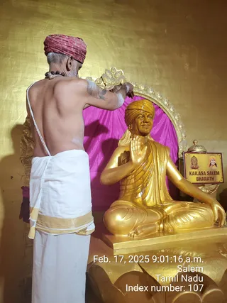 A bare-backed priest in a red-and-gold turban and white dhoti with a gold border stands with his back to the camera, applying white paste or sandal to a golden ornamental frame behind a seated golden statue. The statue, in saffron-gold, wears a golden turban, a draped robe and sits cross-legged with its right hand raised in abhaya mudra on a lotus-shaped base, set against a pink cloth backdrop. A small sign on the right reads KAILASA SALEM BHARATH beside a portrait of a bearded figure in an orange turban. Timestamp reads Feb. 17, 2025 9:01:16 a.m., Salem, Tamil Nadu. Statue is a depiction, not a physical person, so the a person candidate is only a statue.