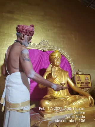A photograph inside a temple-like room with a gold-painted wall. A bare-chested priest in a white dhoti with a gold border and a red turban, with rudraksha beads at his neck, stands at left performing a ritual. He holds a small oil lamp, and white paste marks his arm. Before him sits a large gold-painted seated statue, with a gold turban, an upraised hand and a gold robe, set against a magenta cloth and an ornate gold frame. A small yellow sign reads KAILASA SALEM BHARATH. The timestamp reads Feb. 17, 2025, 9:00:16 a.m., Salem, Tamil Nadu.
