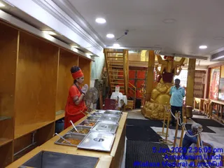 Interior photo of a temple dining space during Annadanam lunch service. A woman in orange robes and an orange head wrap with a long plait stands behind a counter of steel serving trays holding curries, wearing transparent gloves and facing left. Beside her, a man in a light teal shirt stands near a large golden statue of a seated deity with raised arms and a red cloth, and an older man in a blue shirt crouches at a brass rope barrier. Gold-framed portraits hang on the right, with a gold staircase and red-painted doors behind. Overlaid blue timestamp reads 5 Jan 2026 2:36:05 pm, Annadanam Lunch.