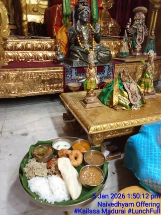 A photograph of a naivedhyam food offering placed on the marble floor in front of a gilded wooden platform. A round metal thali sits on a banana leaf holding white rice, a flatbread, two fried vadas, a sambar-style dish, coconut chutney, a pickle, a red-orange curry and a brown gravy, with a small piece of banana leaf visible. Behind it, on the gold-carved platform, stand small brass and green-clad deity murti, one with a dark-green silk cloth draped over it and a jewelled necklace. A seated dark metal deity murti with raised hand and rudraksha-style beads sits in the background beside gilded panels and a red cloth. A blue woven cloth edge appears at the right. The overlay reads 5 Jan 2026 1:50:12 pm, Naivedhyam Offering. a person is not visibly present.
