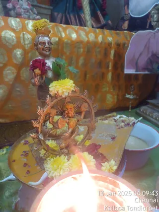 A close photo of a Nataraja murti, a bronze-toned dancing Shiva within a circular arch of flame, decorated with yellow chrysanthemums and red rose petals on a brass tray. Beside it stands a smiling bronze devotee figure holding a red rose and leafy stems, and a white enamel bowl of milky liquid sits to the right. A glowing oil lamp in the foreground, an orange and gold brocade cloth behind, and a small brass lamp on the right complete the abhishekam offering setup. A date and location stamp reads 13 Jan 2025 04:09:43 Chennai Kailasa Tondiorpet. No people are visible.