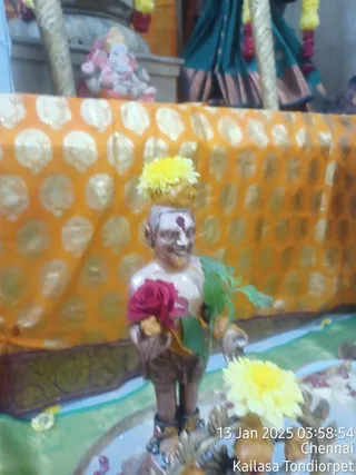 A blurry photograph of a small painted figurine of a standing devotional figure with a yellow marigold on its head, wearing a red flower garland draped over its shoulder, holding green leaves in one hand. The figure is set in front of an orange and gold patterned silk backdrop, with a teal and gold-bordered sari-like drape and red and yellow flower garlands visible above, and a pink and white Ganesha figure in the upper left. Yellow marigold flowers and petals are scattered on a green and white decorated base in the foreground. A timestamp reads 13 Jan 2025 03:58:54 with Chennai and Kailasa Tondiorpet overlaid in the lower right. No living people are visible.