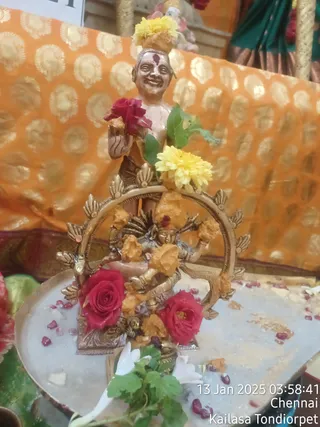 A photograph of a small brass Nataraja-style murti of Shiva as the cosmic dancer, standing within a decorative arched brass prabhavali on a silver tray. The figure is coated with orange-yellow turmeric or sandalwood paste and decorated with red roses, yellow marigolds and green leaves. A standing figure of a smiling devotee-like statue with a red tilak and yellow flower on the head rises above the arch, and red rose petals and seeds are scattered on the tray. The background is an orange cloth with gold circular motifs. A timestamp overlay reads 13 Jan 2025 03:58:41 Chennai Kailasa Tondiorpet. No living people are visible.