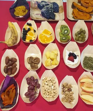 Overhead photo of a food offering spread on a red tablecloth. Small wooden leaf-shaped boat plates hold sliced and whole fruits: a banana, blueberries and orange segments, kiwi slices, green olives, dates, dried figs, pineapple chunks, a yogurt dollop topped with berries, pumpkin seeds, purple grapes, cashews, walnuts and apple slices. Larger square wooden trays at the top hold roasted purple and orange sweet potatoes and sliced pumpkin. No people are visible in the frame.