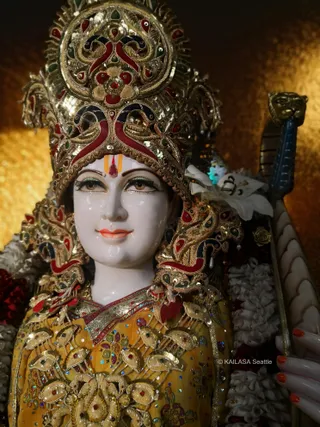 Close-up photograph of a white marble deity murti with an ornate gold and red jeweled crown and a red-and-gold tilak on the forehead. The murti wears a mustard-yellow embroidered garment with gold zari work and jeweled motifs, and white jasmine-style garlands hang at its side. A brass-headed staff with a blue shaft rests beside it against a glowing golden background. Lilies and flower petals are tucked near the shoulder. No physical people are visible.
