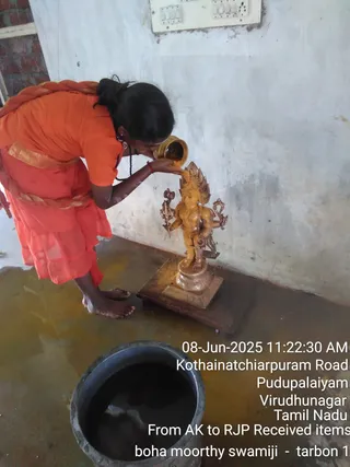 A barefoot person in an orange sari with a gold-bordered waist wrap pours liquid from a golden vessel over a small bronze deity murti with multiple arms, standing on a wooden base against a pale wall. A dark metal pot of liquid sits in the foreground on a wet floor. A timestamp and location overlay appears in the lower right corner.