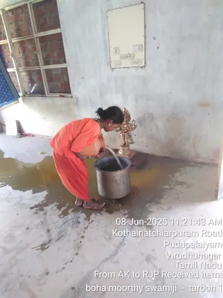 A photograph of a person in an orange sannyasi-style outfit bending over a steel vessel marked A15, pouring water from a copper pot into it on a wet concrete floor. A small gold-coloured deity murti stands beside the vessel against a plain wall, with a window and a blue cloth at the left edge. The person has hair tied back and is barefoot. Timestamped text in the lower right gives a Kothainatchiarpuram Road, Pudupalaiyam, Virudhunagar, Tamil Nadu location, with a note about items received from AK to RJP. The page refers to a Rajapalayam ritual, but the image shows only a water-pouring scene, so no specific ritual is established.