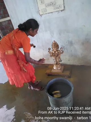 A young woman in an orange top and orange-red skirt with a gold border leans over a gold-coloured metal murti of a multi-armed standing deity on a wooden platform against a plain white wall, holding a small lamp or flame near the figure while a large grey metal vessel of liquid sits on the wet floor in front. A red-banded arm and bare feet are visible; she wears a dark braid and a red bindi-style mark. Water covers the floor. The photo carries a timestamp and caption overlay.