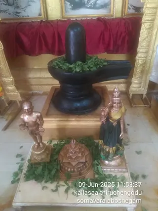 A dark black Shiva lingam rises from a black yoni-peetha base set on a gold-trimmed square platform, decorated with green bilva leaves at its base. Red silk drapes hang behind it, framed by gold carved pillars and two framed black-and-white pictures above. In front stand a copper-coloured standing male figure bare-chested with a dhoti and a copper-coloured female figure in a dark green and yellow sari holding a pot, beside a copper sri yantra meru on a white marble platform scattered with bilva leaves. No living people are visible. The scene resembles a Shiva abhishekam setup with devotional decorations.