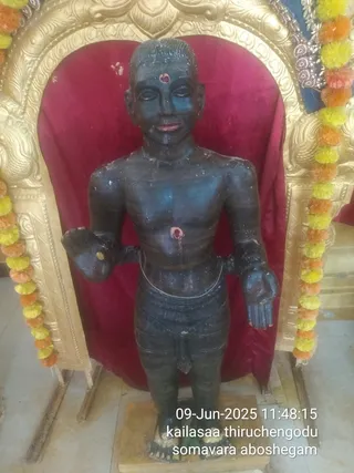 A dark, standing statue of a bare-chested male figure with a red tilak on the forehead, red markings on the chest and a red bindi-like dot at the navel, wearing a short dhoti-style garment and a thin waist chain. The statue stands on a wooden base in front of a deep red velvet backdrop framed by an ornate gold arch. Marigold and yellow-orange flower garlands hang along the sides of the arch. The image is a photograph with an on-screen timestamp and caption overlay reading 09-Jun-2025 11:48:15 and somavara aboshegam, indicating an abhishekam ritual setting. No living people are visible.