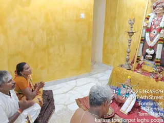 A photograph inside a small room with bright yellow walls and a white marble floor. On the left, a man in a white dhoti with grey hair and a moustache sits with palms pressed together, and beside him a woman in an orange blouse and yellow-patterned sari with red bangles sits with folded hands, gazing upward in prayer. At the lower centre, an elderly bare-chested priest with grey hair sits facing a red-cloth-covered altar. On the right, a deity murti in a red garment is decorated with white and multicoloured flower garlands and a rudraksha-style necklace; a brass oil lamp stands before it, and a brass pot and offerings rest on a yellow cloth. A timestamp and the location Tondiarpet, Chennai, Tamil Nadu are overlaid, with a caption reading kailasa tondiorpet laghu pooja.