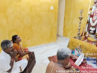A small yellow-walled shrine room with white floor tiles. On the left, a man in a white dhoti and white shirt sits with palms pressed together in prayer, beside a woman in an orange blouse and patterned sari with red bangles, looking up toward the shrine. In the lower centre, a bare-chested older man with grey hair sits near red-cloth offerings and a brass plate. On the right, a tall brass oil lamp stands before a deity murti draped in garlands of orange and white flowers, with a gold-framed backdrop. A timestamp reads 16 Jun 2025 16:49:31 at 278/47 4th Street, Tondiarpet, Chennai, Tamil Nadu.