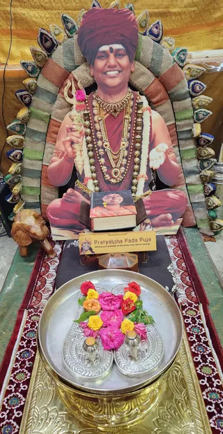 A life-size cardboard cutout of a smiling man in a maroon turban and maroon robe sits cross-legged before a multicoloured beaded backrest, holding a silver trident adorned with a pink flower in one hand and an open palm in the other, wearing layered gold necklaces, rudraksha-style beads and a red forehead dot. In front, a book titled Living Enlightenment rests on a wooden stand, and a wooden sign reads Pratyaksha Pada Puja. A silver plate holds silver padukas, or sandals, surrounded by red, pink, orange and yellow flowers, with two small silver vessels beside them. A carved wooden elephant stands at left and a gold-and-red patterned cloth covers the platform. This is a depiction on a cutout, not a person in the scene.