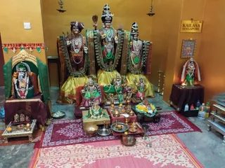Indoor temple shrine with orange walls and a grey floor, showing a group of deity murtis dressed in gold and green silk with green and gold garlands. The central standing figure wears a tall black and gold crown and holds a fan-like staff, flanked by two standing figures with turbans and jewelled necklaces. A seated figure in a green-draped niche sits on the left beside a Happy Diwali banner. Offerings are arranged on the floor, including a plate of marigolds, oil lamps, a kalasha and brass vessels on red mats. A sign on the wall reads KAILASA SINGAPORE and a framed painting hangs on the right. No living people are visible.