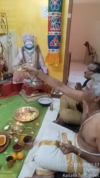 A photo of a small puja inside a yellow-walled room at a KAILASA centre. Two men sit on a white cloth with green mat before a cutout portrait of a turbaned figure with a long beard, draped in ornate necklaces, set on a red-draped stand. A priest in a white dhoti with gold border, bare-chested and wearing a gold necklace, raises a small bowl or lamp toward the image, while a second man in khaki trousers sits behind with hands folded in namaskar. Brass and copper vessels, a silver plate with flowers, oranges, red kumkum and a trishul-like object sit on the green mat. A yellow-and-orange banner with elephant and lotus motifs hangs beside the figure, and a tridented murti stands at the left edge. A timestamp reads 10 Mar 2025 08:52:37 Chennai, Kailasa Tondiorpet.