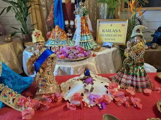 A photograph of a shrine table at the KAILASA Guadeloupe Goyave centre, with no living people visible. A gold-framed sign reads KAILASA GUADELOUPE GOYAVE. Two small statues in ornate gold and green garments stand on the left and right. Behind them, a larger deity murti wears a garland of purple and white flowers and a blue and gold robe. A brass tray holds pink and purple flower petals, and a small seated figure with a rudraksha mala sits on a white lace cloth scattered with pink and purple flower petals. Brass bowls and a palm plant complete the setting.