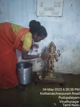 A photograph of a woman bending over a small brass deity murti on a low wooden stand against a pale wall. She wears a red saree with a yellow blouse, bangles and a drop earring, and holds a small spoon or ladle near the figure. Beside the stand sit small brass bowls and a large steel pot on the floor. An electrical switch board is mounted on the wall above. A timestamp overlay reads 04-May-2025 6:30:38 PM, Kothainatchiarpuram Road, Pudupalaiyam, Virudhunagar, Tamil Nadu.