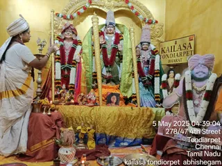 Interior shrine photo at KAILASA Tondiarpet with a golden arched backdrop and a sign reading KAILASA TONDIARPET BHARAT. Three large deity murtis stand under flower garlands: the central figure wears a tall crown and green cloth, flanked by a figure in red-and-gold turban and a figure in a tall ornate crown and blue-green dress. A woman in a white and gold-bordered saree and white headcloth stands at left holding a tall lamp-like ritual object near the murtis. Brass oil lamps, a small Ganesha figure, a framed portrait, and offering plates sit on a gold cloth-covered altar. At right a cutout board shows a smiling man in a pink-and-gold turban with a garland and rudraksha-style beads, with a photo caption and timestamp overlay reading 5 May 2025 04:39:04 278/47 4th Street Tondiarpet Chennai Tamil Nadu.