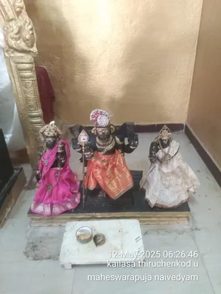 Indoor photo of three dark-stone or black-painted deity murtis on a black pedestal before a gold-toned wall and carved gold frame. The central figure wears an orange and gold silk dhoti, a jeweled crown, and holds a trident-like staff and other attributes. The figure to the left wears a magenta silk sari with gold border and a jeweled headdress; the figure to the right wears a cream and gold patterned sari with a crown. A small white stone platform in front holds a brass bowl and a coconut-shaped offering with naivedyam. A timestamp overlay reads 12-May-2025 06:26:46 and the caption text reads kailasa thiruchenkod u maheswarapuja naivedyam. No people are visible.