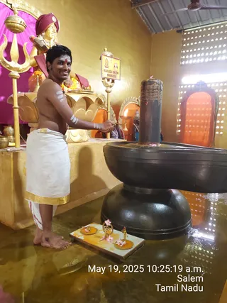 A bare-chested man in a white dhoti with a gold border stands beside a large black lingam on a dark stone yoni-pitha base, pouring or tending it with his hands. He has white ash stripes on his arms and a red tilak on his forehead. Behind him a golden statue of a seated figure wears a maroon turban and saffron-orange garments with garlands, beside a golden trident. A small oil lamp burns on a golden stand, and pink flowers lie on a yellow tray with small brass figures near the base. A sign reads KAILASA SALEM BHARATH. The room has gold-painted walls, a pink curtain and a perforated window. The lower corner carries a timestamp reading May 19, 2025 10:25:19 a.m., Salem, Tamil Nadu.