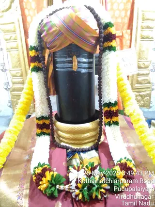 A photograph of a black stone Shivalingam on a golden yoni-pitha base, decorated for worship. The lingam is draped with a multicoloured striped cloth in pink, orange and green, and wrapped with white and yellow marigold garlands with green and maroon floral accents. A long rudraksha mala hangs around it, and a bright yellow-orange tilak mark is painted on the lingam's front. Below, a red and gold silk cloth covers the base with floral offerings. Gold-toned ornamental panels stand behind. A timestamp and location caption in the lower right read 26 May 2025, Kothainatchiarpuram Road, Pudupalaiyam, Virudhunagar, Tamil Nadu. No people are visible.