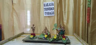 A photograph of a small shrine arrangement set on a dark wooden tray against a gold silk curtain. A paper sign on the curtain reads KAILASA TRINIDAD & TOBAGO. In the centre stands a tiered golden shrivelled-pyramid form, a shivalingam-style base, flanked by a small standing gold figure on the left and a gold deity murti on the right, the right figure seated with a green cloth draped over its lap. Each figure and the central form is decorated with fresh flower garlands in red, yellow and blue-violet blossoms and green leaves. A red and green garland hangs at the right edge of the frame. No people are visible.