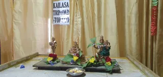 A photograph of a small altar arrangement set against gold silk curtains. A printed white sign reading KAILASA TRINIDAD & TOBAGO is taped to the curtain at the top centre. On a dark wooden platform stands a bronze deity murti seated with a green cloth draped over its lap, beside a small bronze standing figure and a bronze stepped pyramid-shaped object. Red and yellow flowers, blue blossoms and green leaves decorate the platform and base, with a steel bowl of white sweets or offerings in front. A string of red and green decorations hangs at the far right. No people are visible.