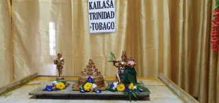 Photograph of a small altar set on a dark wooden platform against a gold satin drape. A paper sign reading KAILASA TRINIDAD & TOBAGO is pinned to the drape at top centre. Three gold-coloured murti stand on the platform: a small standing figure of a devotee at left, a tiered stepped shrine-form pyramid in the centre, and a seated deity with a raised hand holding a pink lotus and a companion figure at right. Blue and yellow flowers, pink lotus blooms and green leaves are arranged around the base of each figure. A red floral garland hangs at the upper right edge of the frame. No people are visible.