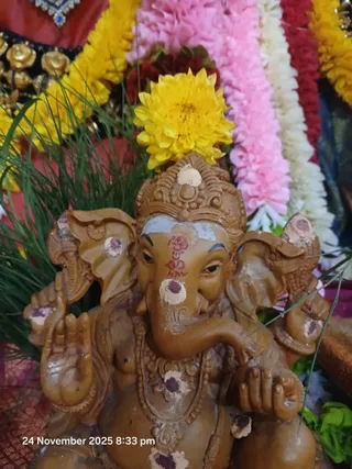 Close-up photograph of a brown clay murti of Lord Ganesha, seated with an upraised hand holding a sweet and his trunk curled toward the mouth, with a crown and ornate necklace. Dabs of white and purple kumkum or vibhuti mark the forehead, trunk and ears, and a yellow marigold sits atop the head. Garlands of yellow, pink, white and red flowers and green grass surround the deity. The frame has a timestamp reading 24 November 2025 8:33 pm. No people are visible.